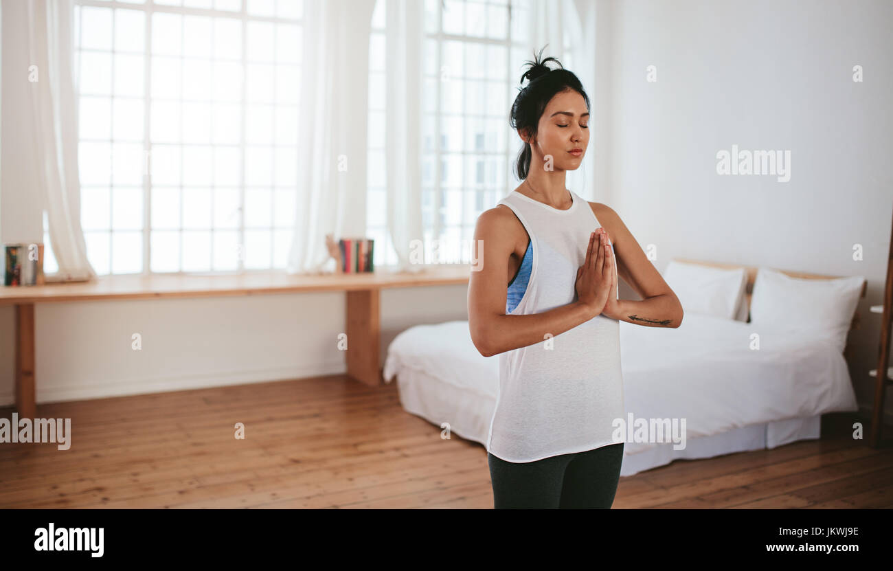 Portrait of healthy young woman standing with her hands joined and eyes closed at home. Fitness female meditating indoors. Stock Photo