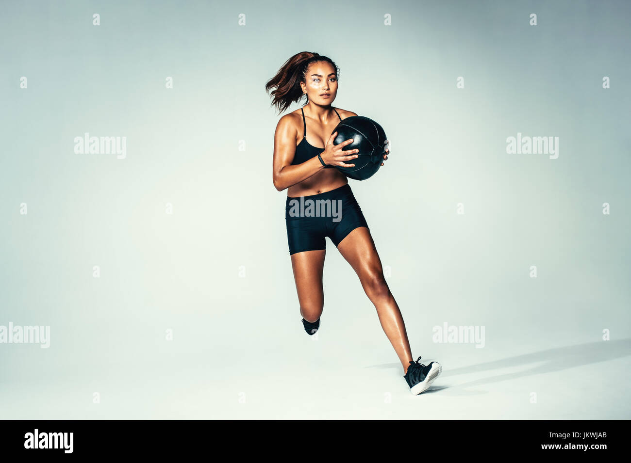 Studio shot of fit young woman running with medicine ball. Female model with muscular body working out with fitness ball over grey background. Stock Photo