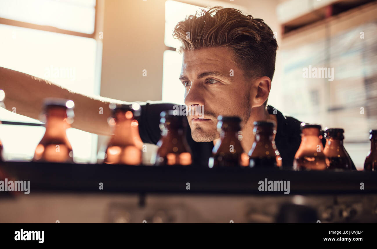 Shot of young man examining the beer bottles on conveyor at brewery factory. Male brewer supervising the process of beer manufacturing in factory. Stock Photo