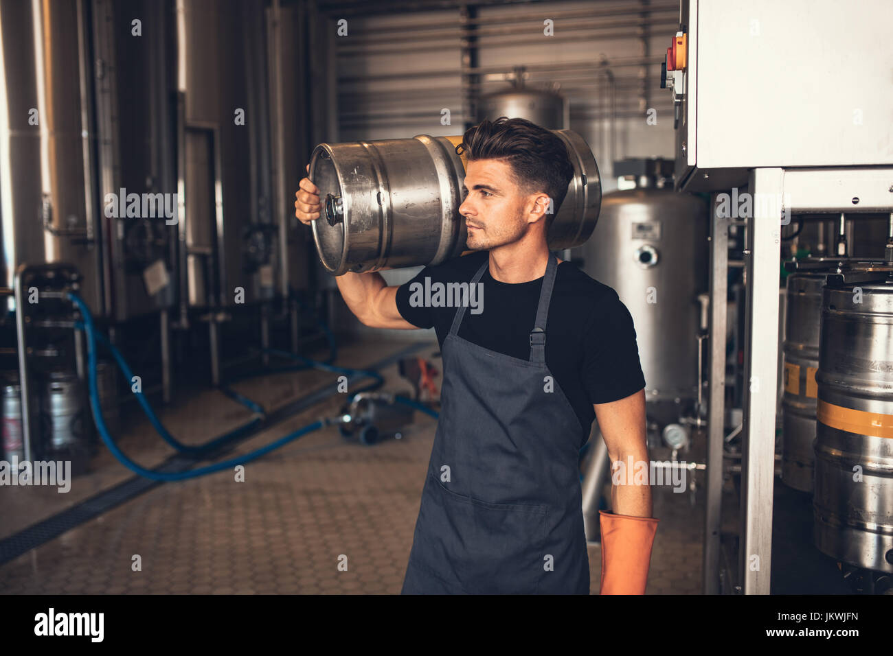 Young male brewer carrying keg at brewery. Manual worker with metal beer barrel craft beer manufacturing plant. Stock Photo