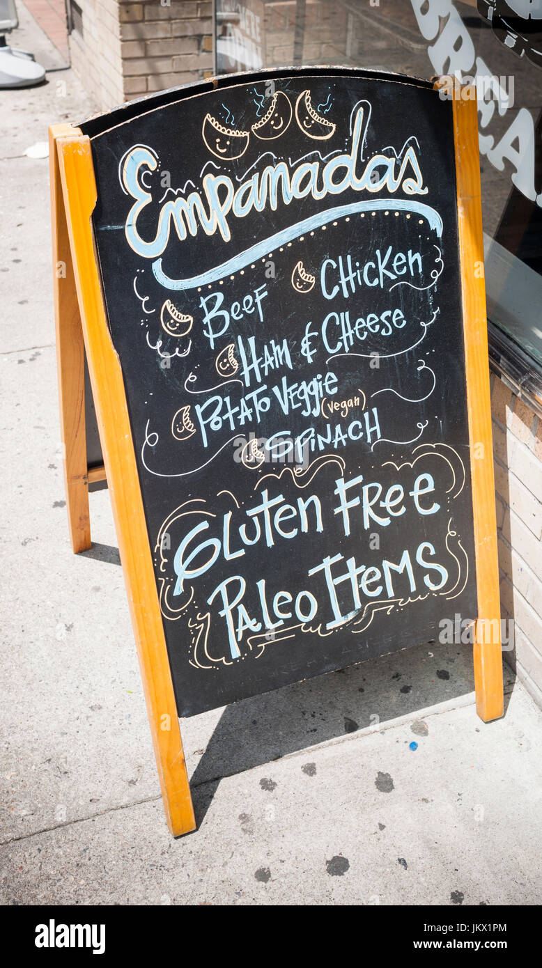 A sign outside a restaurant in New York on Monday, July 17,  2017 promote their gluten free and paleo menu items as well as their empanadas. The protein gluten is found in wheat and is a flavoring and thickening food additive. People with celiac disease, dermatitis herpetiformis  and wheat allergies eat a gluten-free diet to prevent their disease or allergy. (© Richard B. Levine) Stock Photo
