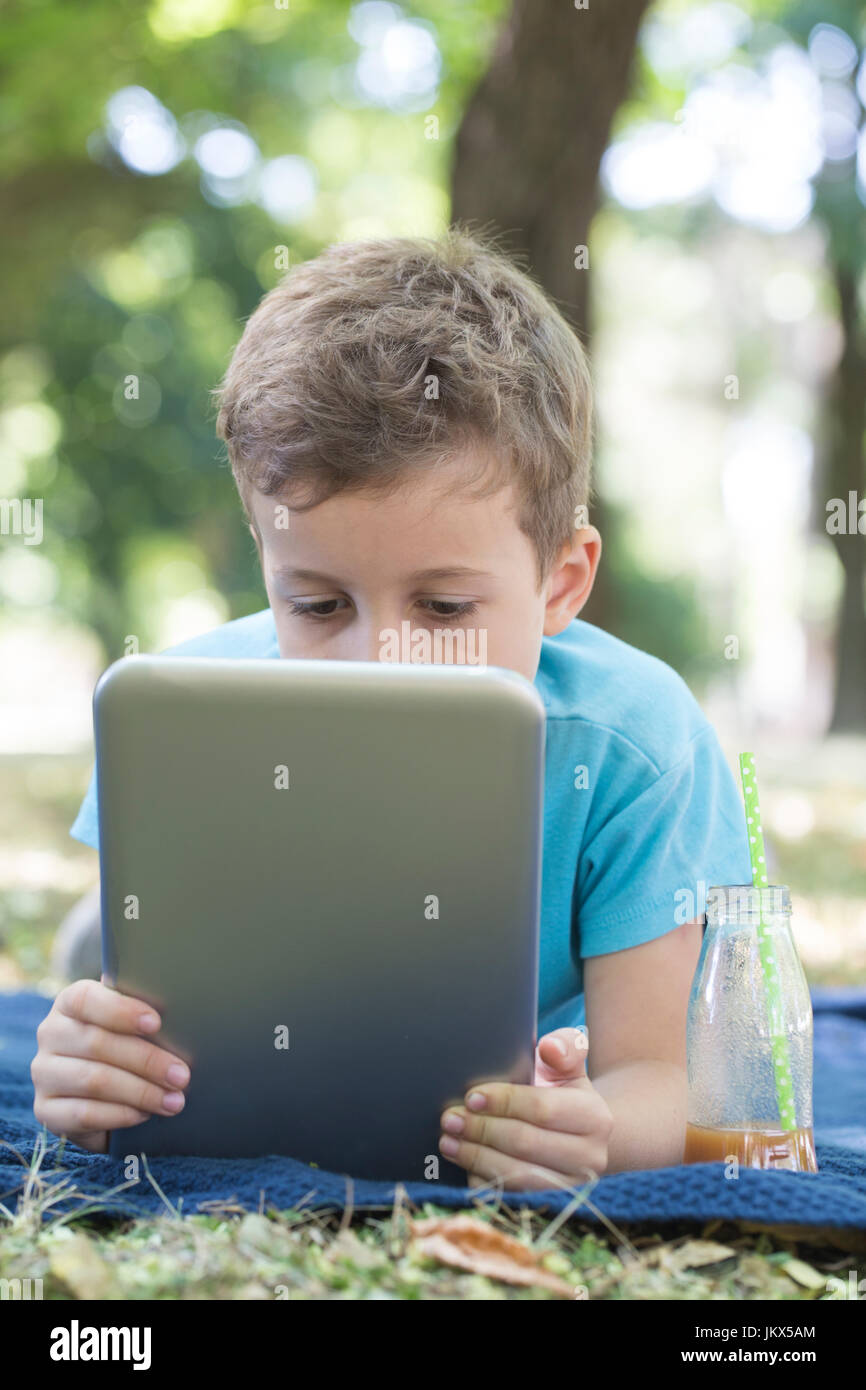 Little cute boy lying on the grass and looking carefully at his tablet ...