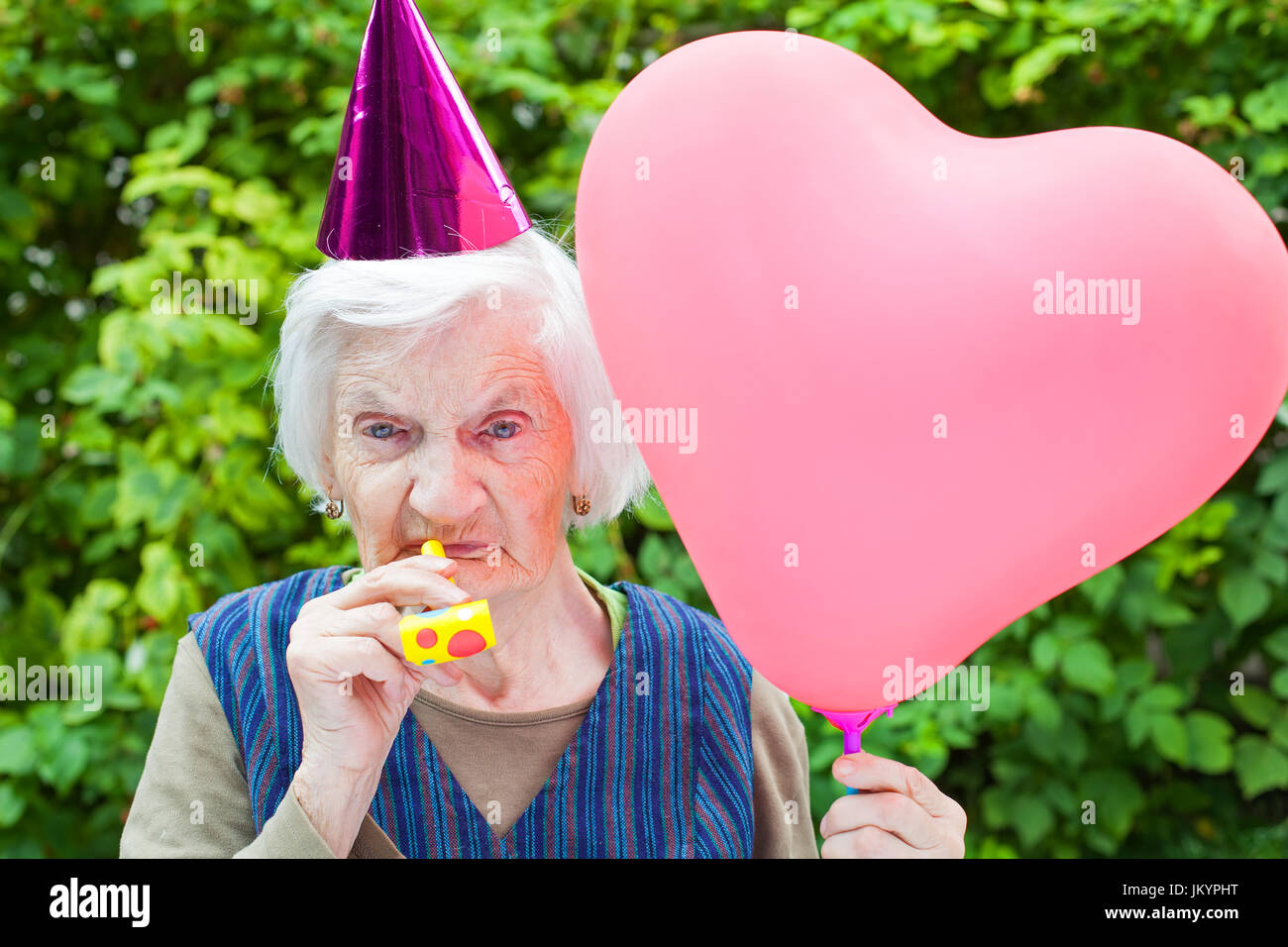 Picture of a happy elderly woman celebrating birthday holding a heart shaped balloon an blowing a whistle outdoor Stock Photo