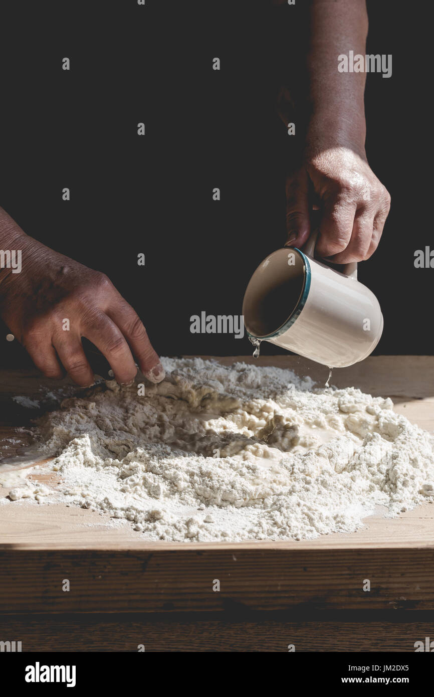 Senior woman kneads pastry, pouring water from mug to flour. Vertical crop, subdued colors, details on old working hands and food ingredients Stock Photo