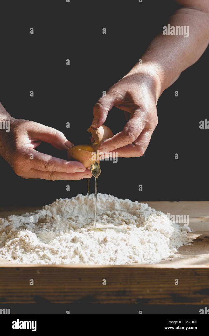 Senior woman kneads pastry, adding egg to flour. Vertical crop, saturated, subdued colors, details on old working hands and food ingredients Stock Photo