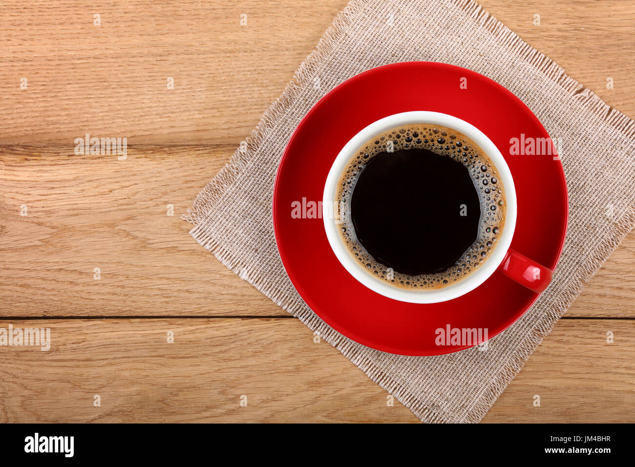 Full big cup of black instant coffee with froth on red porcelain saucer over wooden table with textile tablecloth napkin, close up, elevated top view, Stock Photo