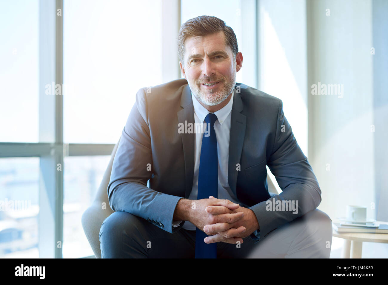 Portrait of a handsome mature business CEO smiling warmly at the camera while sitting in a modern office space with large windows and gentle sunflare Stock Photo