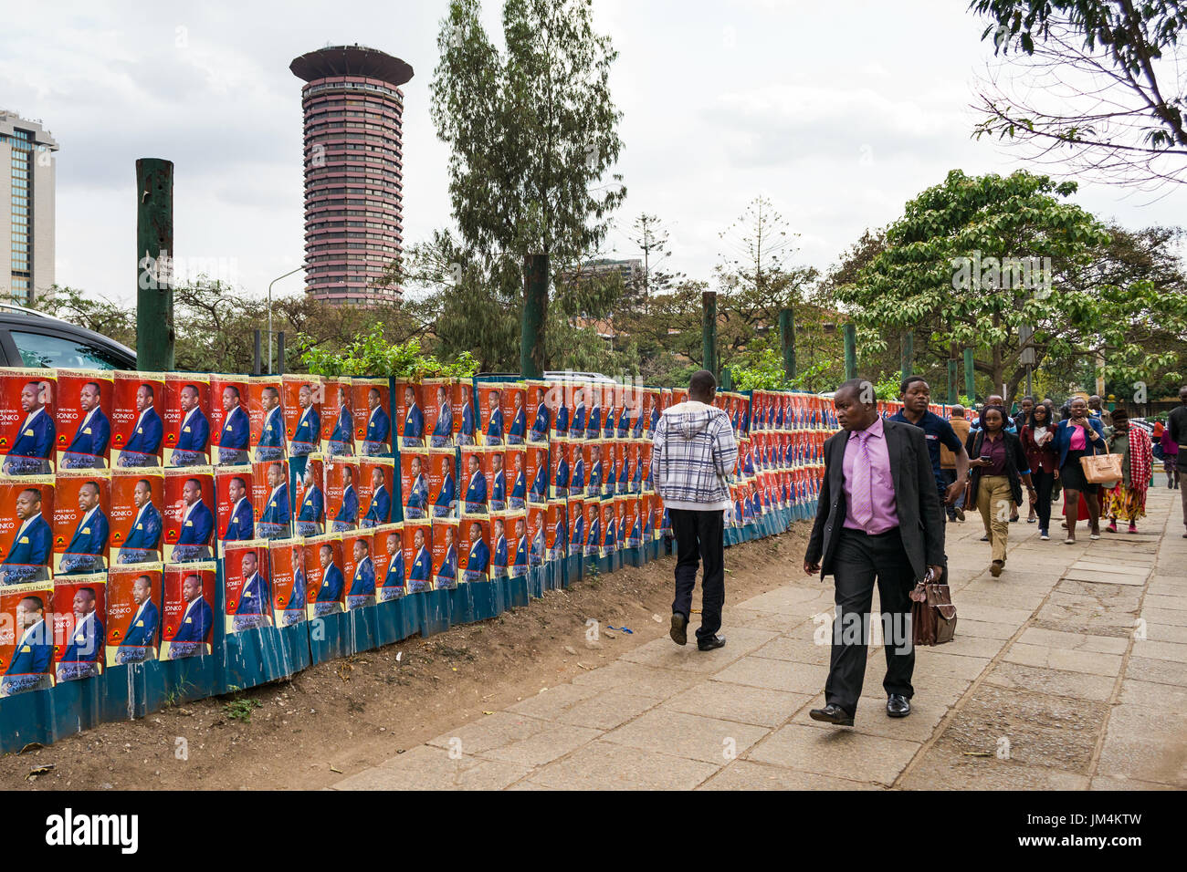 People Walking Past Kenyan Election Candidate Posters On Wall In Nairobi City With Kenyatta International Convention Centre In Background, Kenya Stock Photo