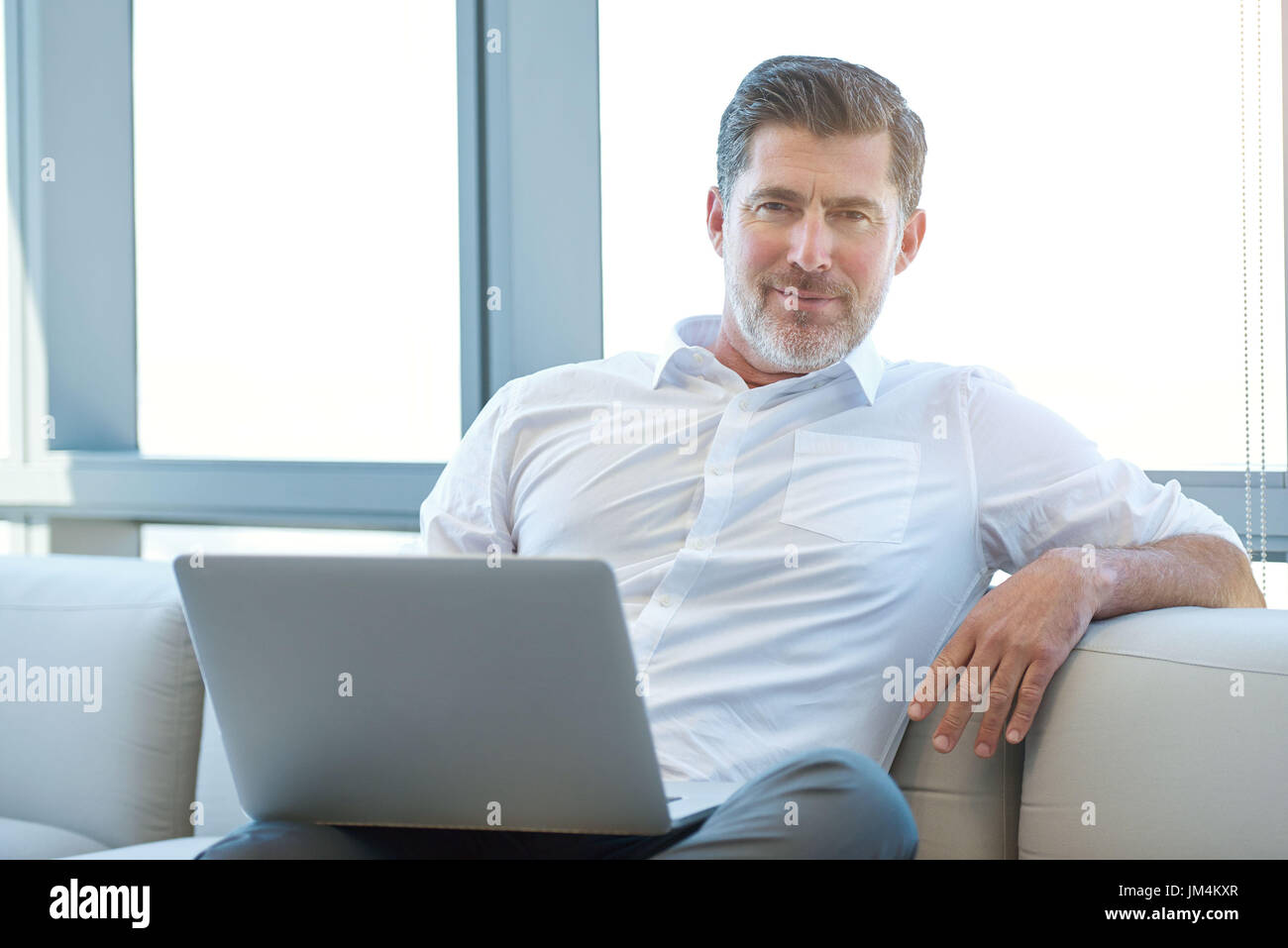 Portrait of a handsome mature businessman relaxing on a couch with a laptop computer, looking positively and confidently at the camera Stock Photo