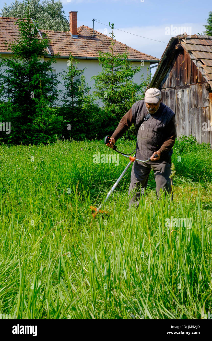 local man using a strimmer to cut long grass in the garden of a rural house in a village in zala county hungary Stock Photo