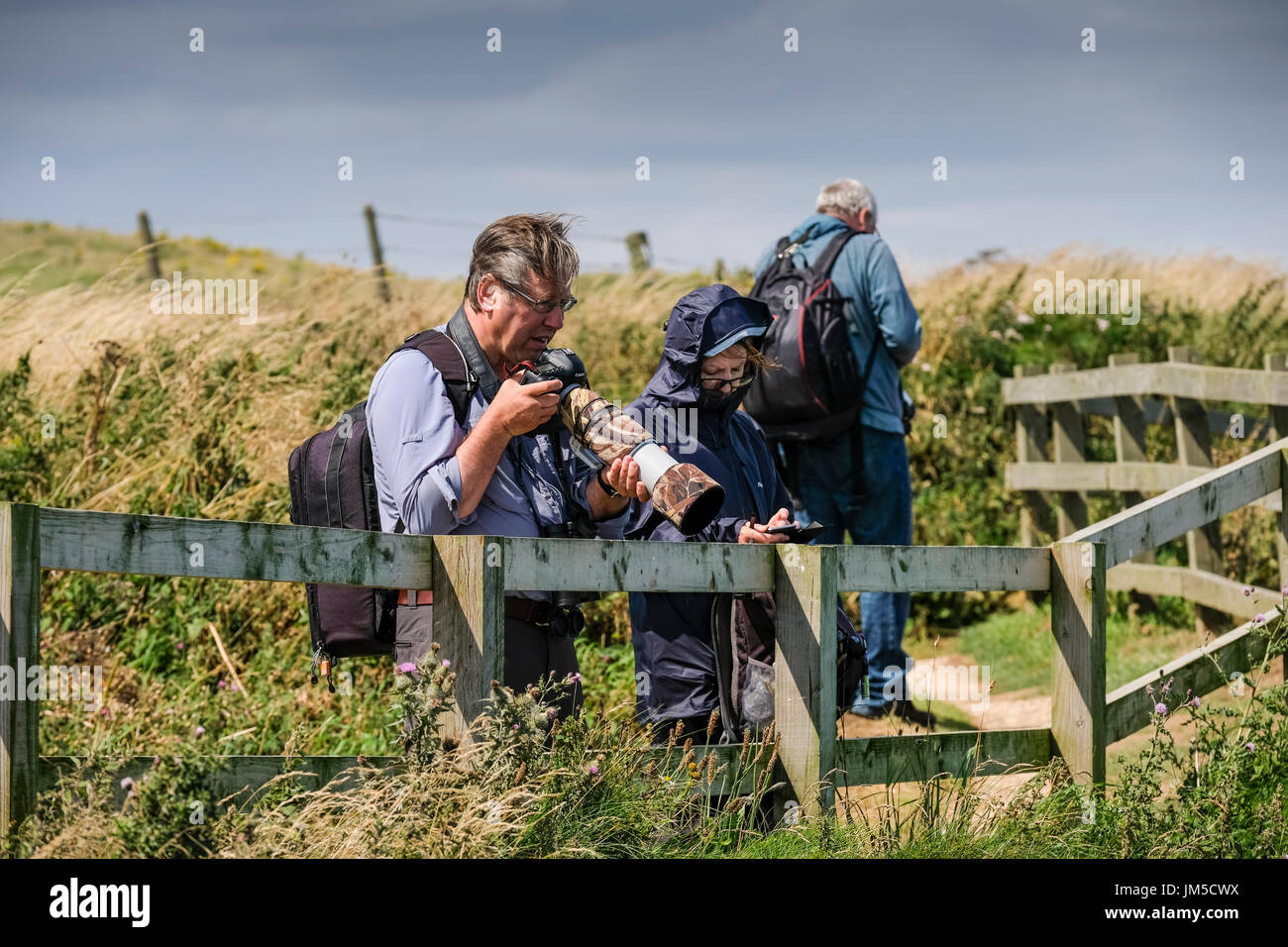 A man standing stood using a camera with a long lens telephoto for bird photography. A female companion woman is stood beside him. Stock Photo