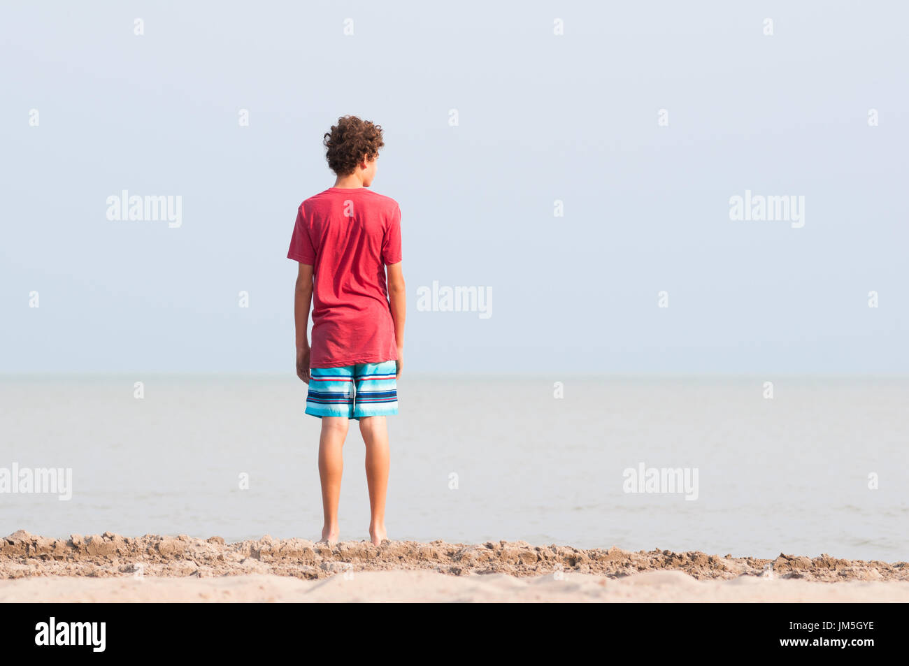 Thirteen year old boy standing on a beach looking out over a lake Stock Photo
