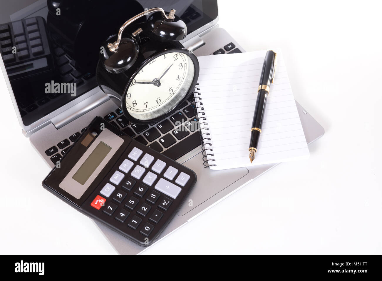 Office objects on laptop keyboard - simple black calculator, blank notebook with fountain pen and classic alarm clock. Viewed from above and isolated  Stock Photo