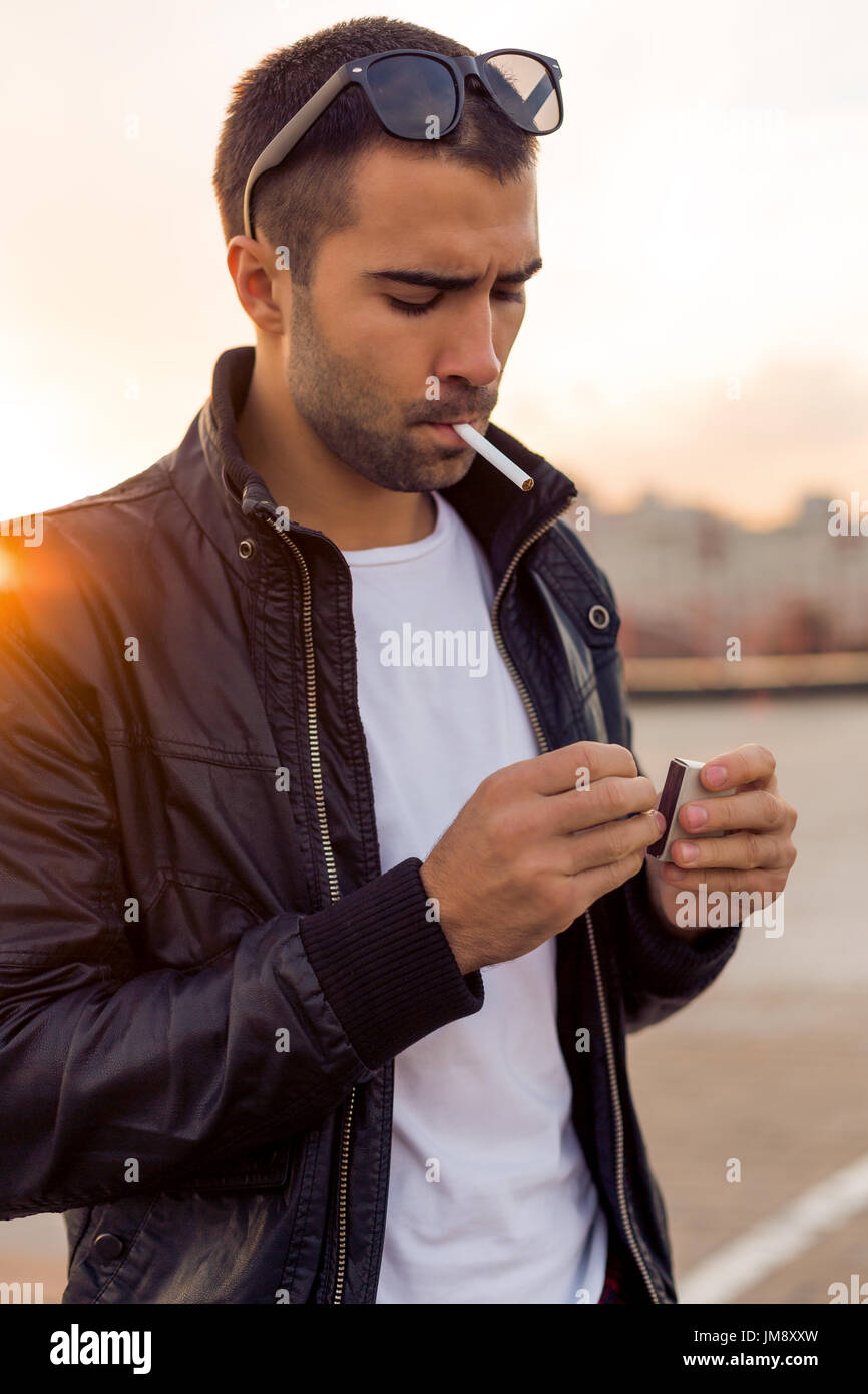 Close up portrait of a handsome rider man with beard and mustache in black  biker jacket, white t-shirt and fashion sunglasses light up cigaret on roof  Stock Photo - Alamy