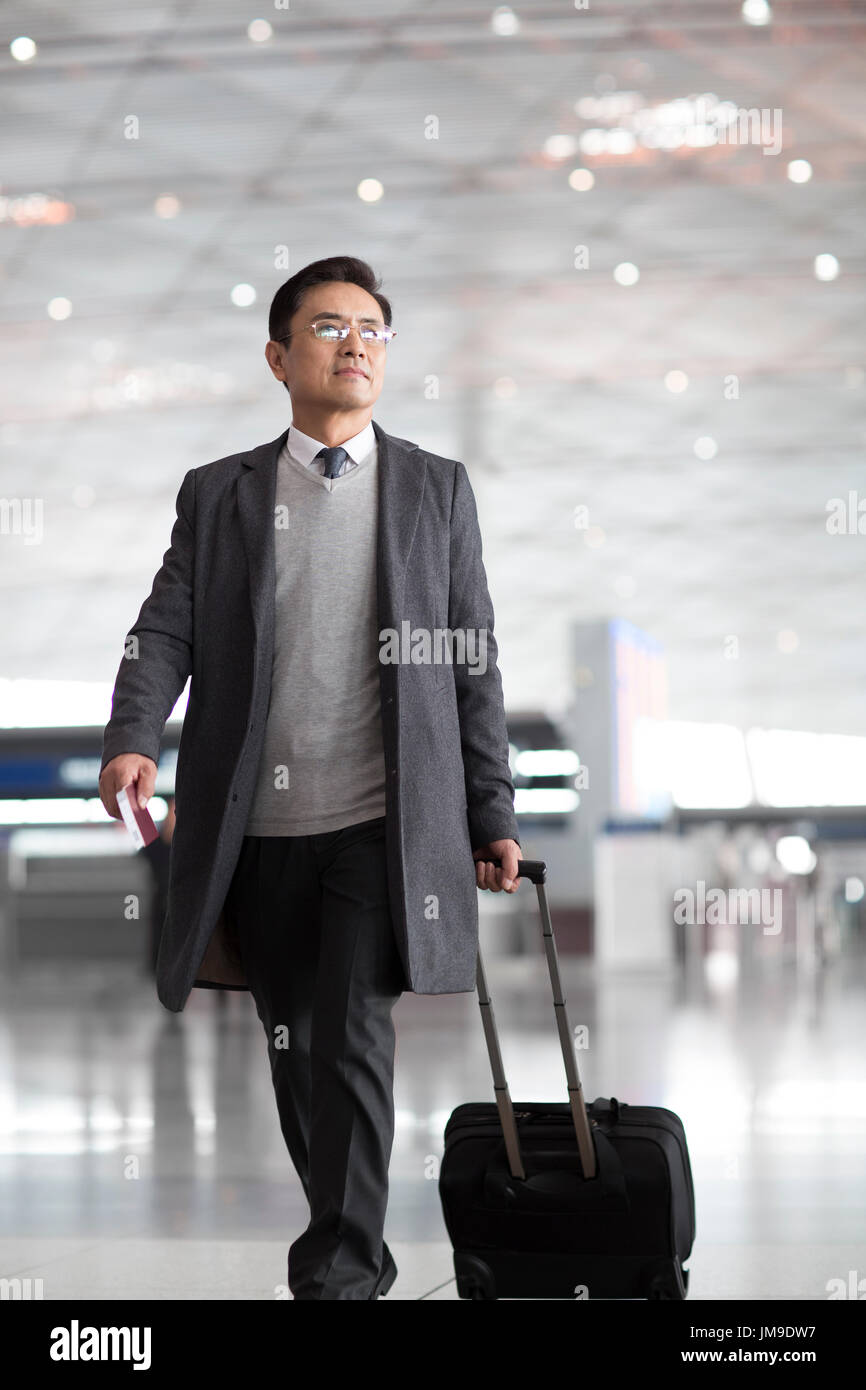 Chinese businessman pulling wheeled luggage in airport lobby Stock