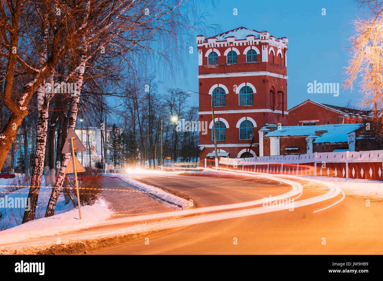 Dobrush, Gomel Region, Belarus. Old Paper Factory Tower At Winter Evening Or Night. Historical Heritage. Lunacharsky Avenue. Traffic Lights Trails On Stock Photo