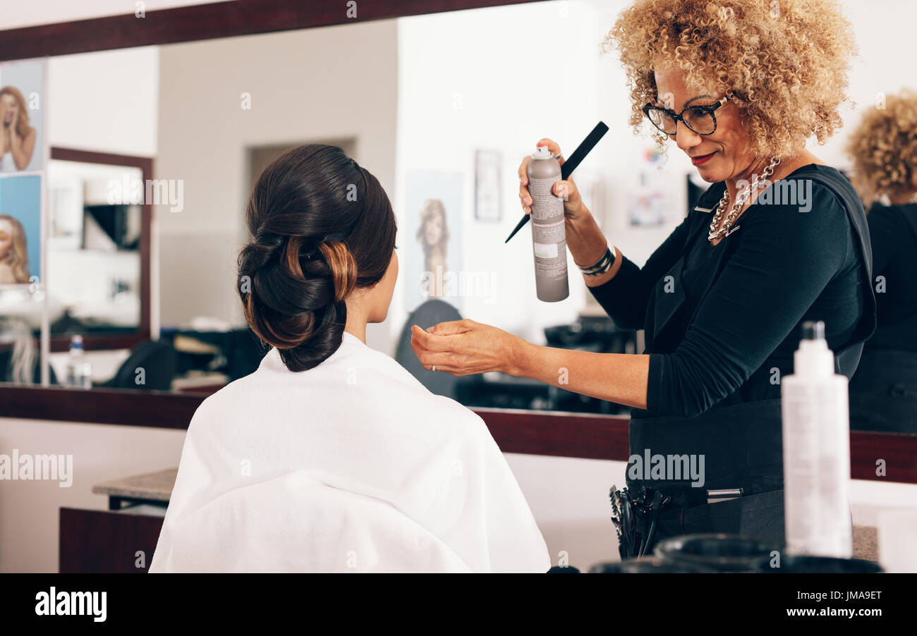 Hairdresser using a hair setting spray for styling the hair. Female hair stylist setting hair in fashionable design at the parlor. Stock Photo