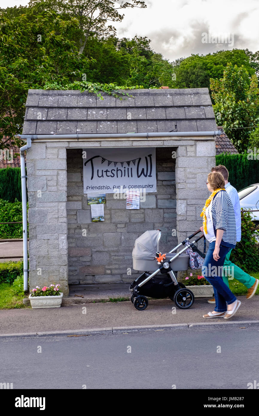 Couple with baby in buggy strolling past community bus stop in Tutshill, Gloucestershire, England UK. Stock Photo