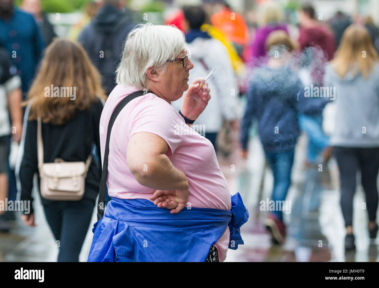 Middle aged overweight Caucasian woman smoking outside in a busy crowded city, in the UK. Unhealthy lifestyle concept. Stock Photo
