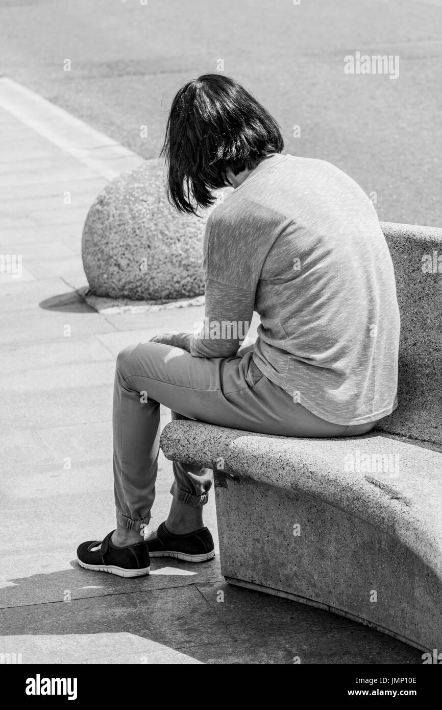 Black and white image of solitary unknown woman seated on bench in sunshine. For loneliness, isolation, separation, rejection, personal space. Stock Photo