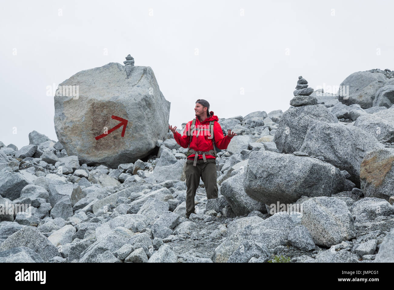 The trail is marked with two large cairns and a giant red arrow painted on a boulder, but this man appears to be lost. Grey, cloudy sky overhead Stock Photo