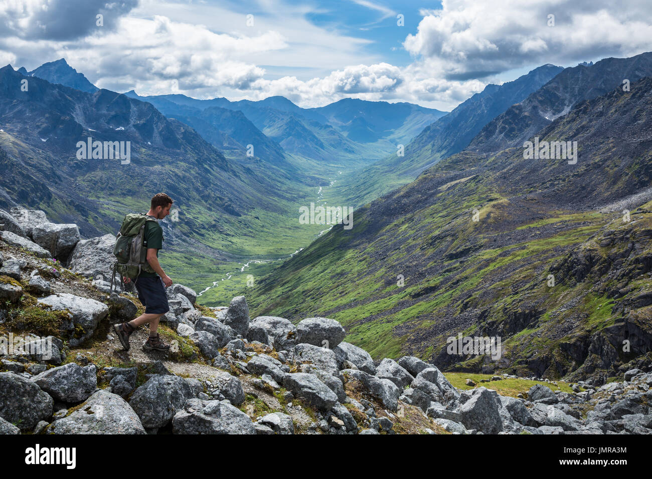 A man in his mid-20's hikes with a daypack down a steep trail overlooking the Little Susitna River in the Gold-Mint Trail Valley of the Hatcher Pass a Stock Photo