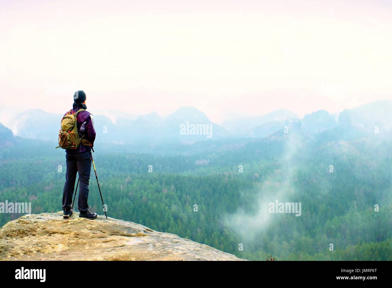 Hiker With Backpack And Poles On Cliff Rock Watching Over The Misty And Foggy Spring Valley To 