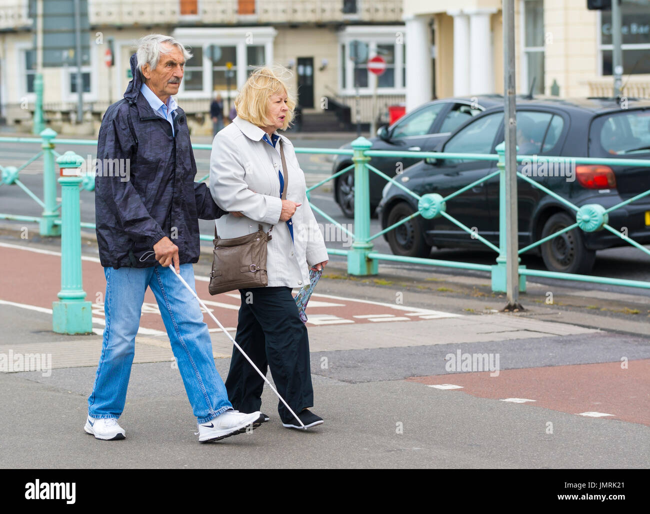 Elderly blind man walking with a white stick with help from a lady. Stock Photo