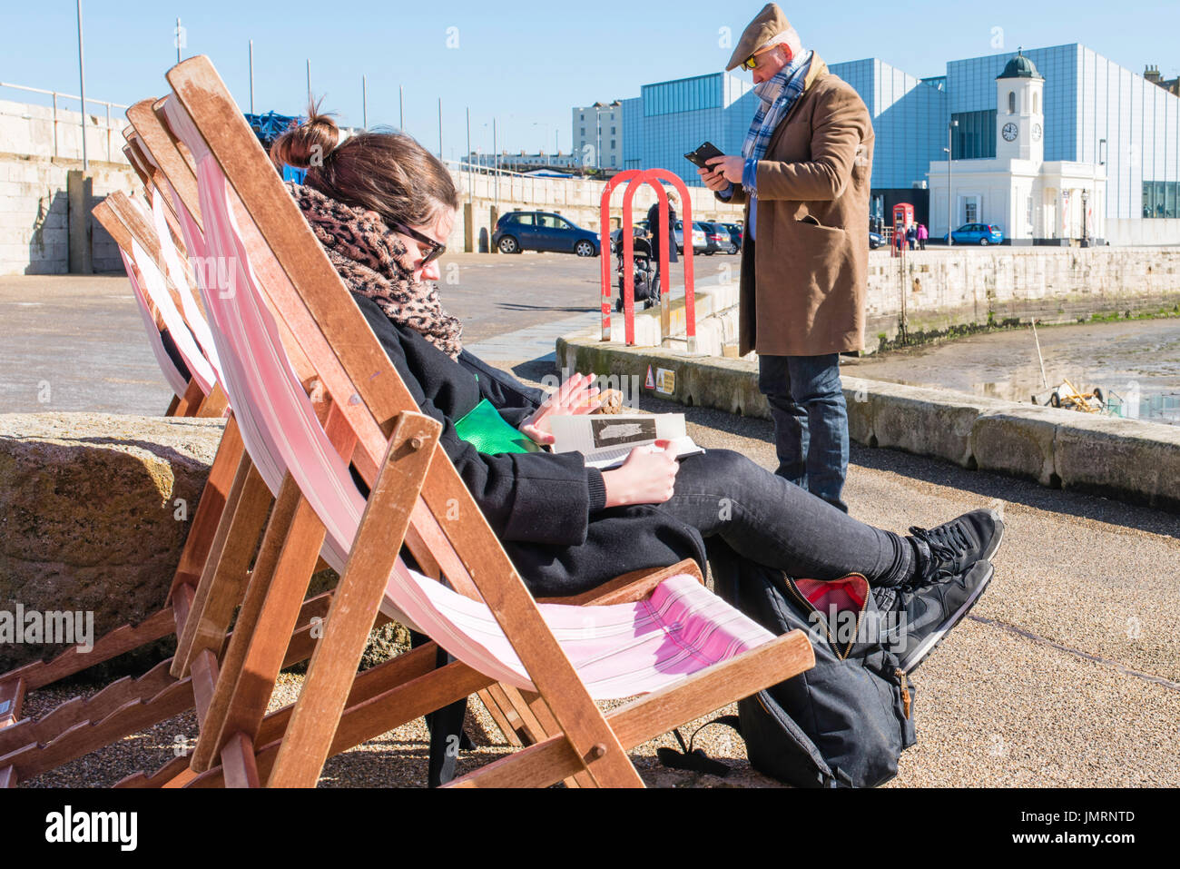 A middle aged man looking at his mobile phone with a young woman reading a book in a deckchair on the harbour wall in Margate, near the Turner gallery Stock Photo