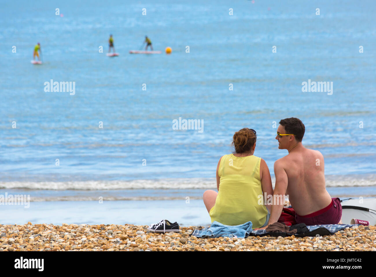 Couple sitting on the beach at Lyme Regis with paddleboarders in the