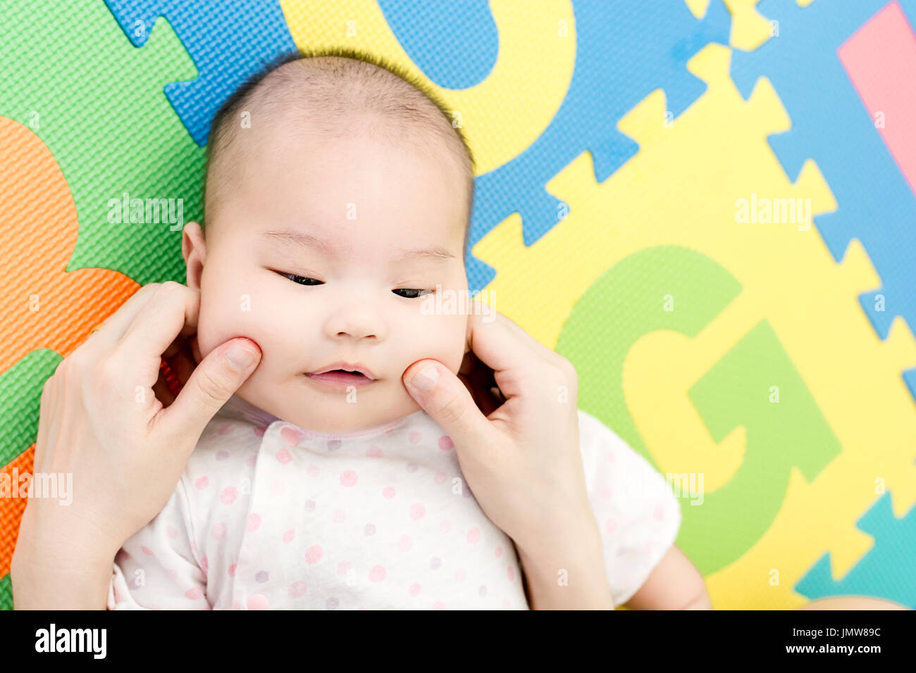 Parent pinch the cheeks a little adorable newborn infant baby girl that lying on the back on colorful eva foam indoors Stock Photo