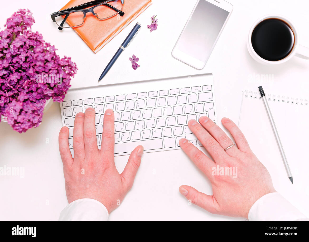 Man fingers working with computer keyboard and workplace with mobile phone, cup of coffee, notebook, pencil, pen, glasses and lilac flowers on the whi Stock Photo