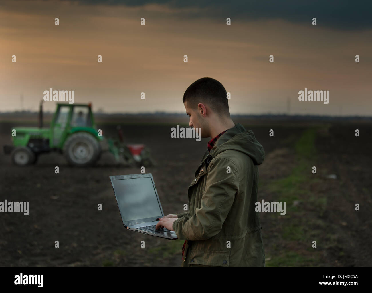 Young farmer with laptop standing in field with tractor harrowing in background Stock Photo