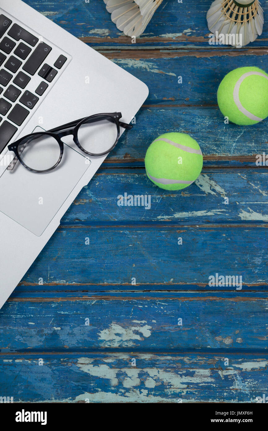 Overhead view of laptop and eyeglasses with shuttlecocks by tennis balls on blue wooden table Stock Photo