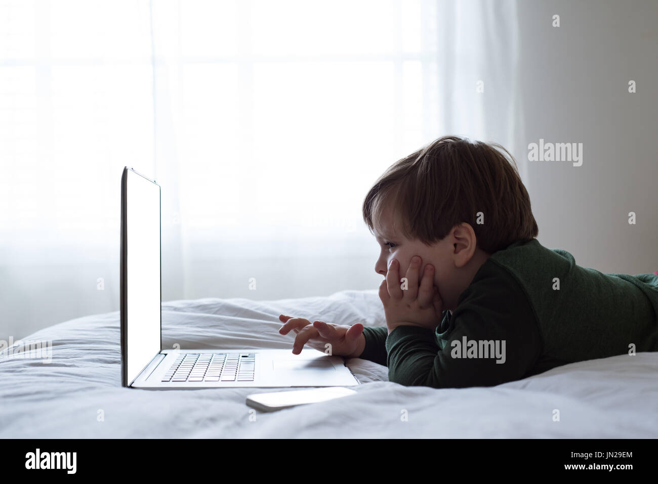 Side view of boy using laptop while lying on bed by window Stock Photo