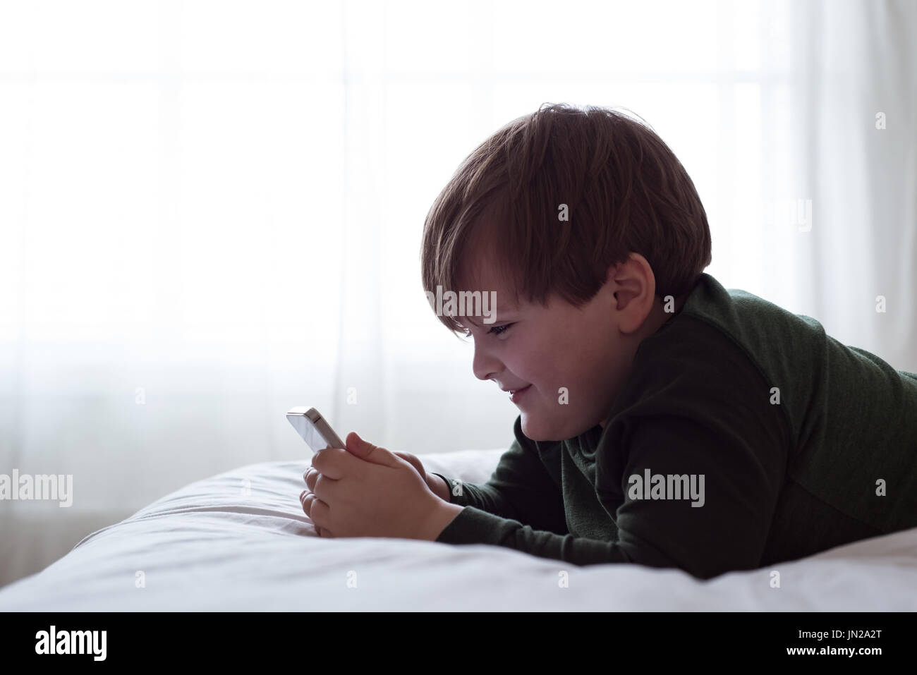 Side view of smiling boy using smart phone while lying on bed by window Stock Photo