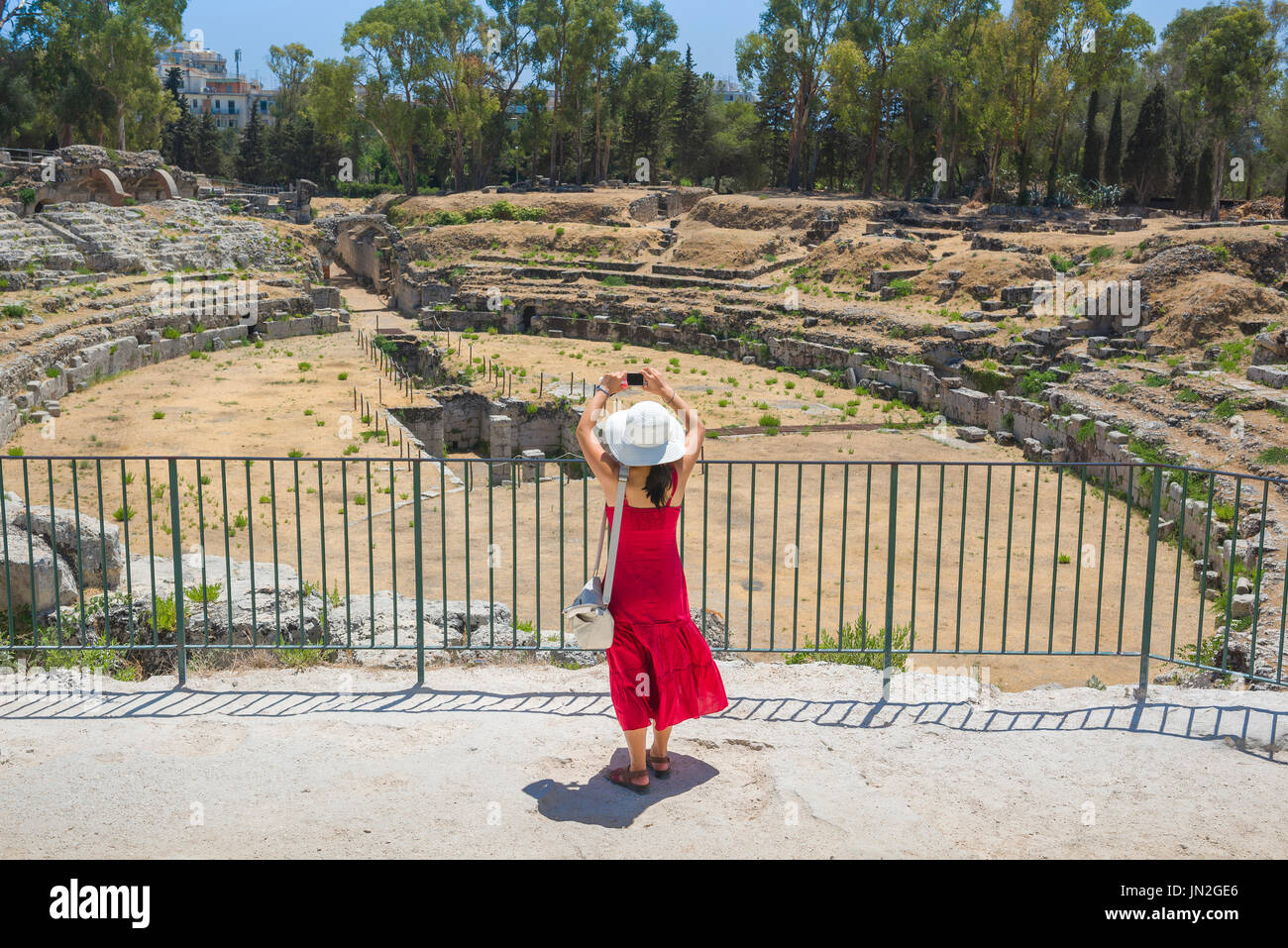 Woman tourist travel concept, view of a young woman in a red dress taking a photo of the Roman ruins in the Archaeological Park in Syracuse, Sicily. Stock Photo