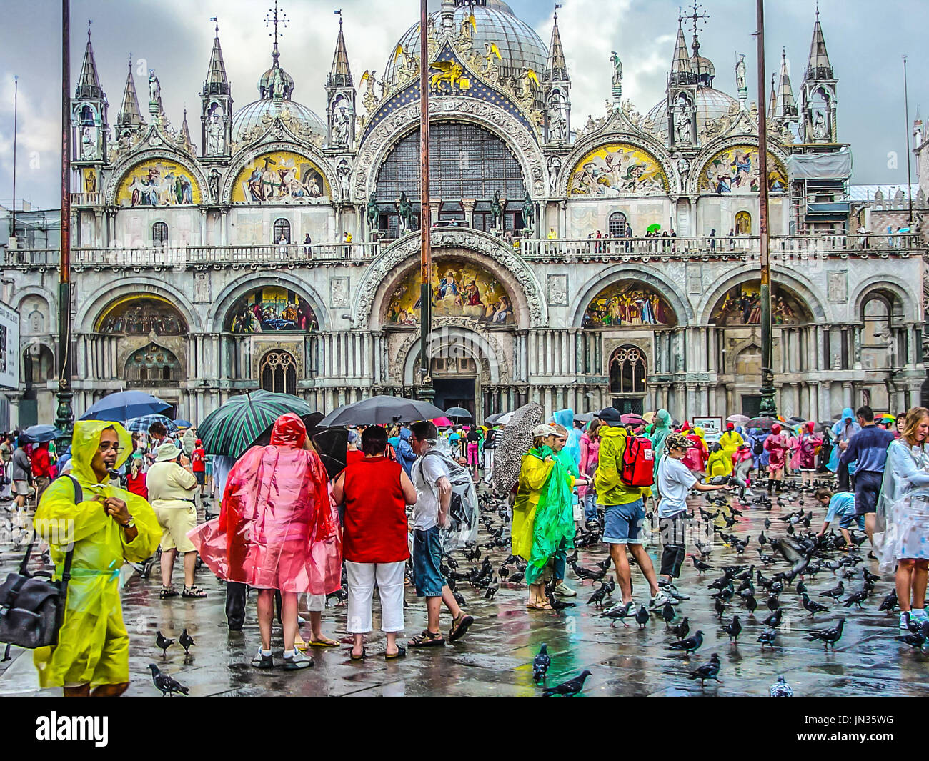 St Mark's cathedral basilica in Venice Italy on a rainy day crowded with tourists in colorful raincoats and umbrellas as pigeons walk around Stock Photo