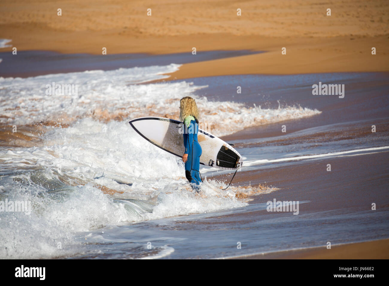 Blonde australian surfer lady in a wetsuit heading to the surf carrying