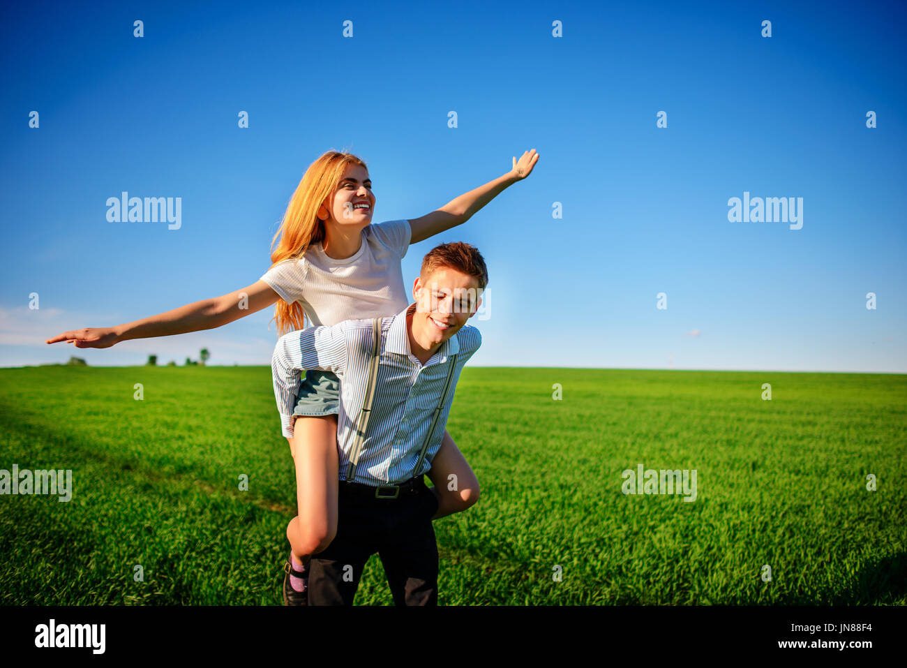 Smiling Man is holding on his back happy woman, who pulls out her arms and simulates a flight against the background of the blue sky and the green fie Stock Photo