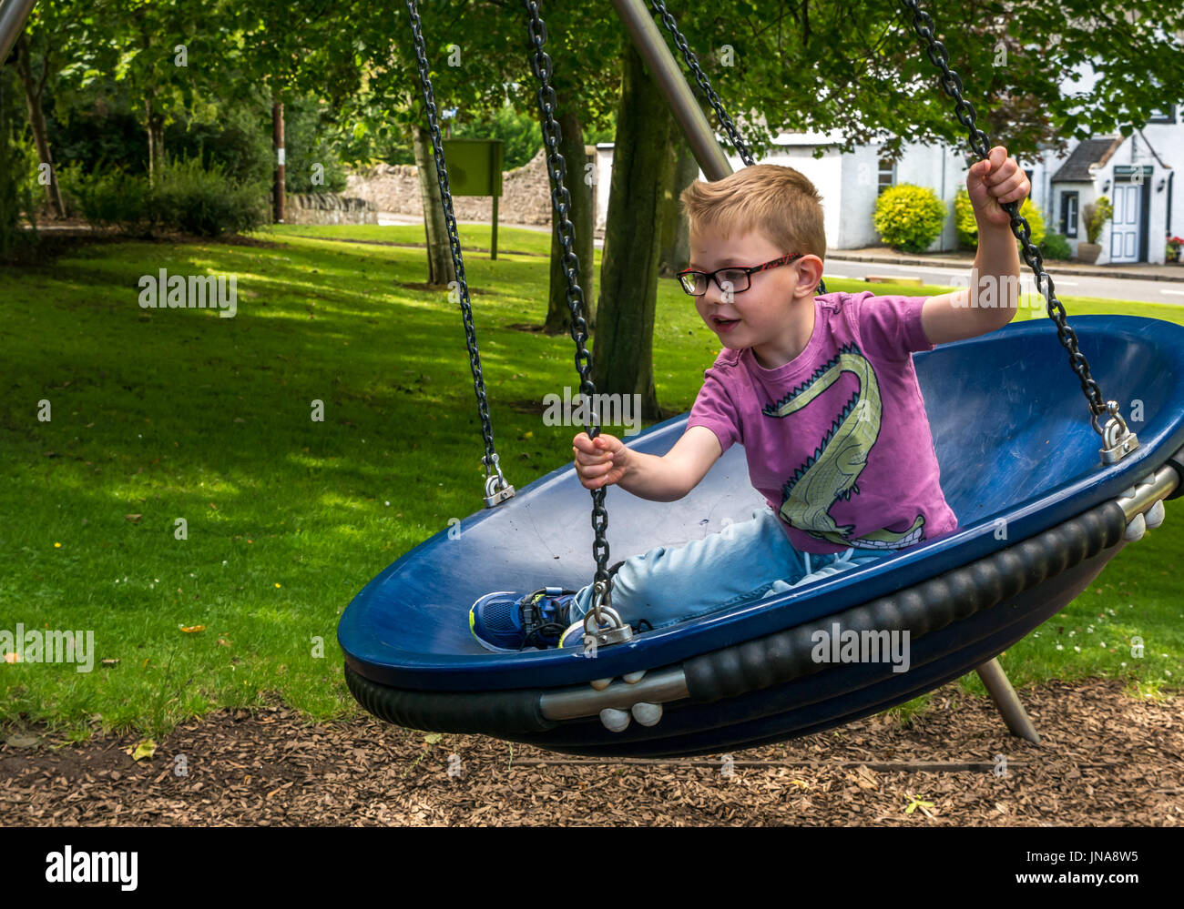 Close up of young boy having fun sitting in circular swing in play park on village green, Dirleton, East Lothian, Scotland, UK Stock Photo