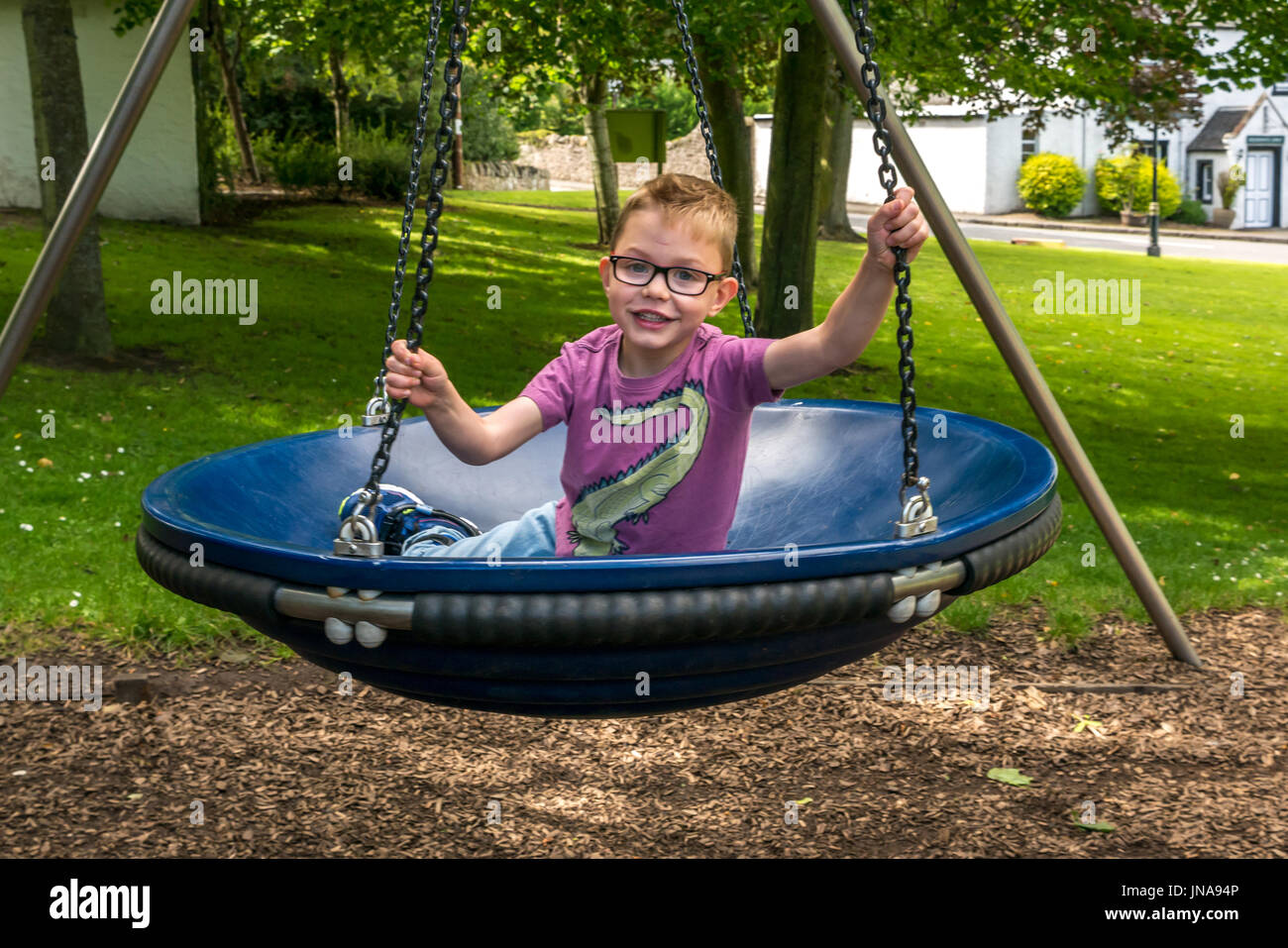 Close up of young boy having fun sitting in circular swing in play park on village green, Dirleton, East Lothian, Scotland, UK Stock Photo