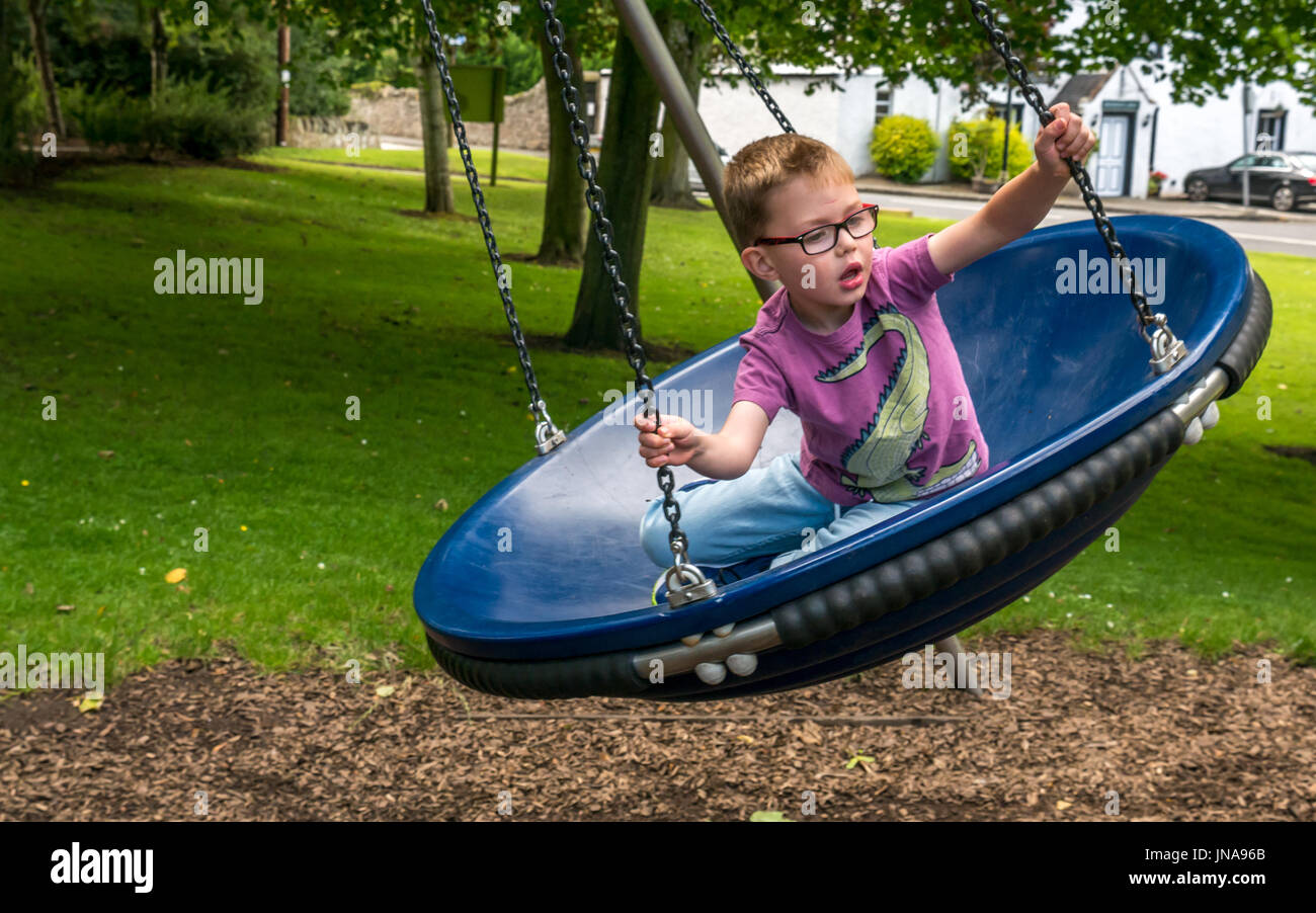 Close up of young boy having fun sitting in circular swing in play park on village green, Dirleton, East Lothian, Scotland, UK Stock Photo
