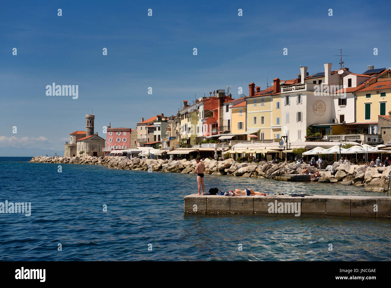 Young couple fishing reading sunbathing on dock at Piran Slovenia on the Adriatic Sea coast with Church of St Clement at Punta Lighthouse Stock Photo