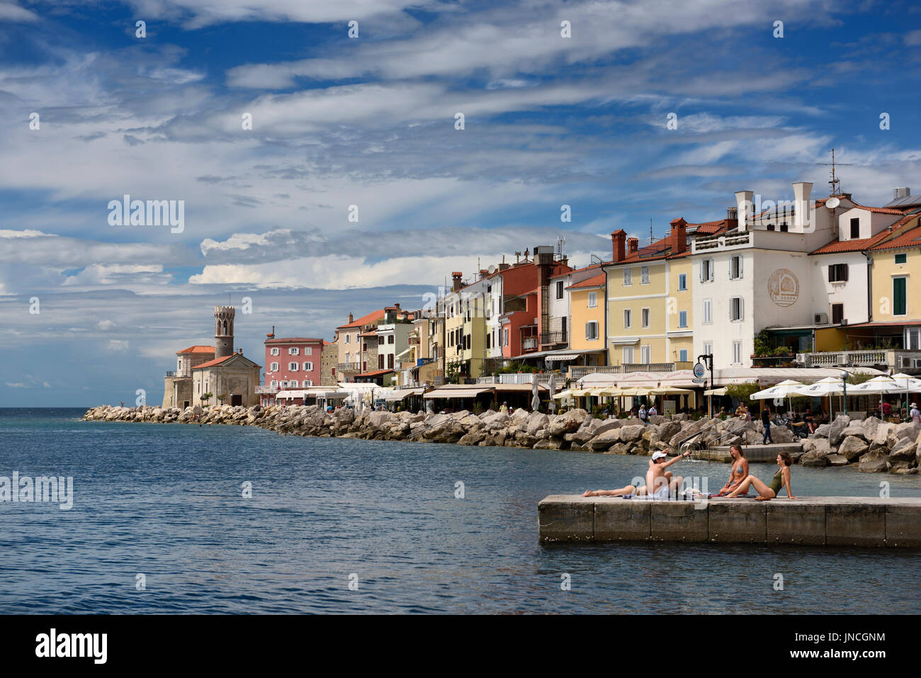 Young people sunbathing on dock at Piran Slovenia on the Adriatic Sea coast with 13th century Church of St Clement at Punta Lighthouse Stock Photo