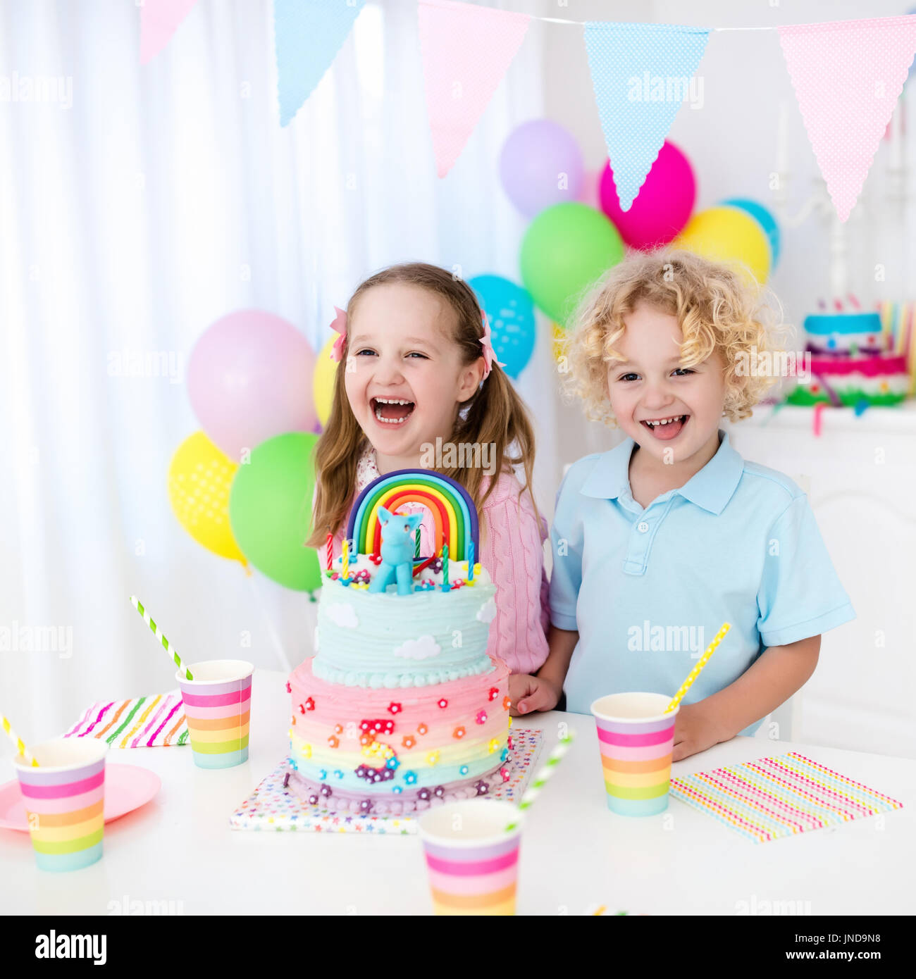 Kids birthday party with colorful pastel decoration and rainbow cake. Girl and boy with sweets, candy and fruit. Balloons and banner at festive decora Stock Photo