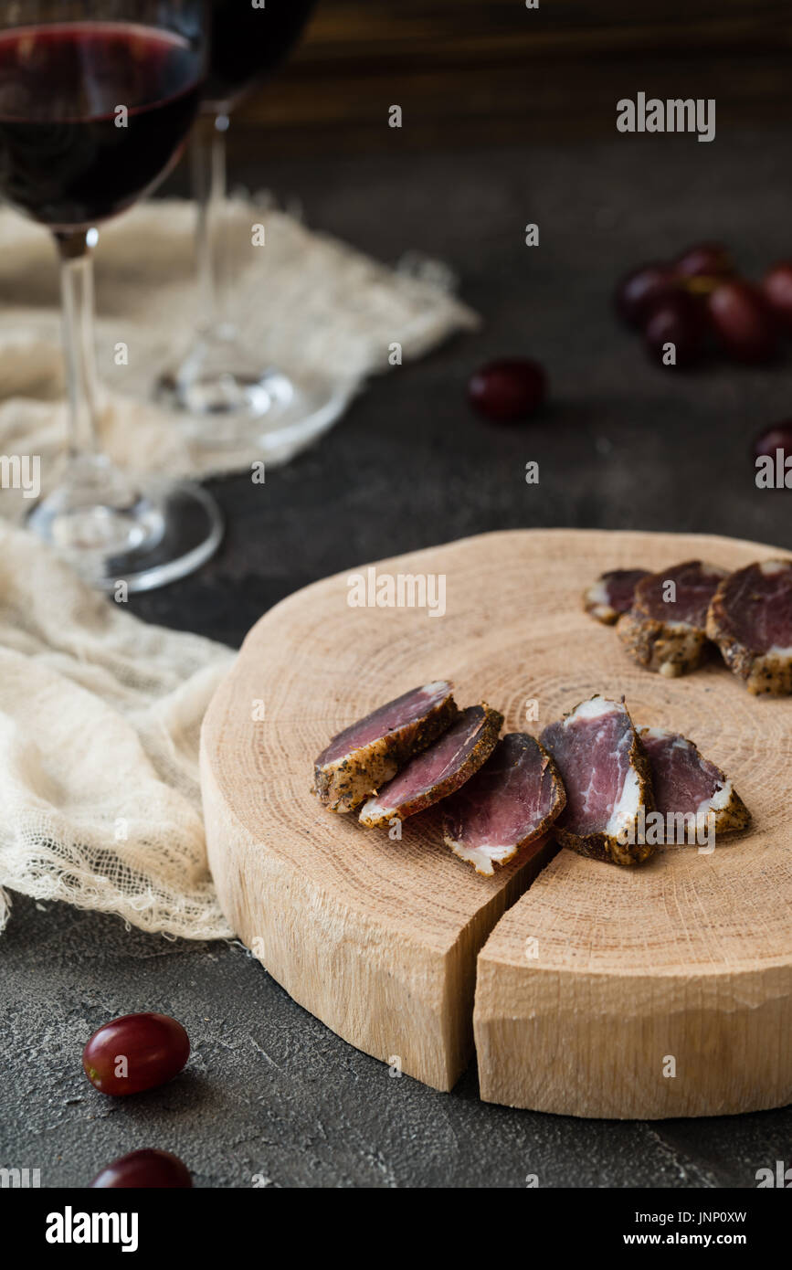 Slices of dried meat on wooden cut, red grapes and two glasses of red wine on dark rustic background Stock Photo