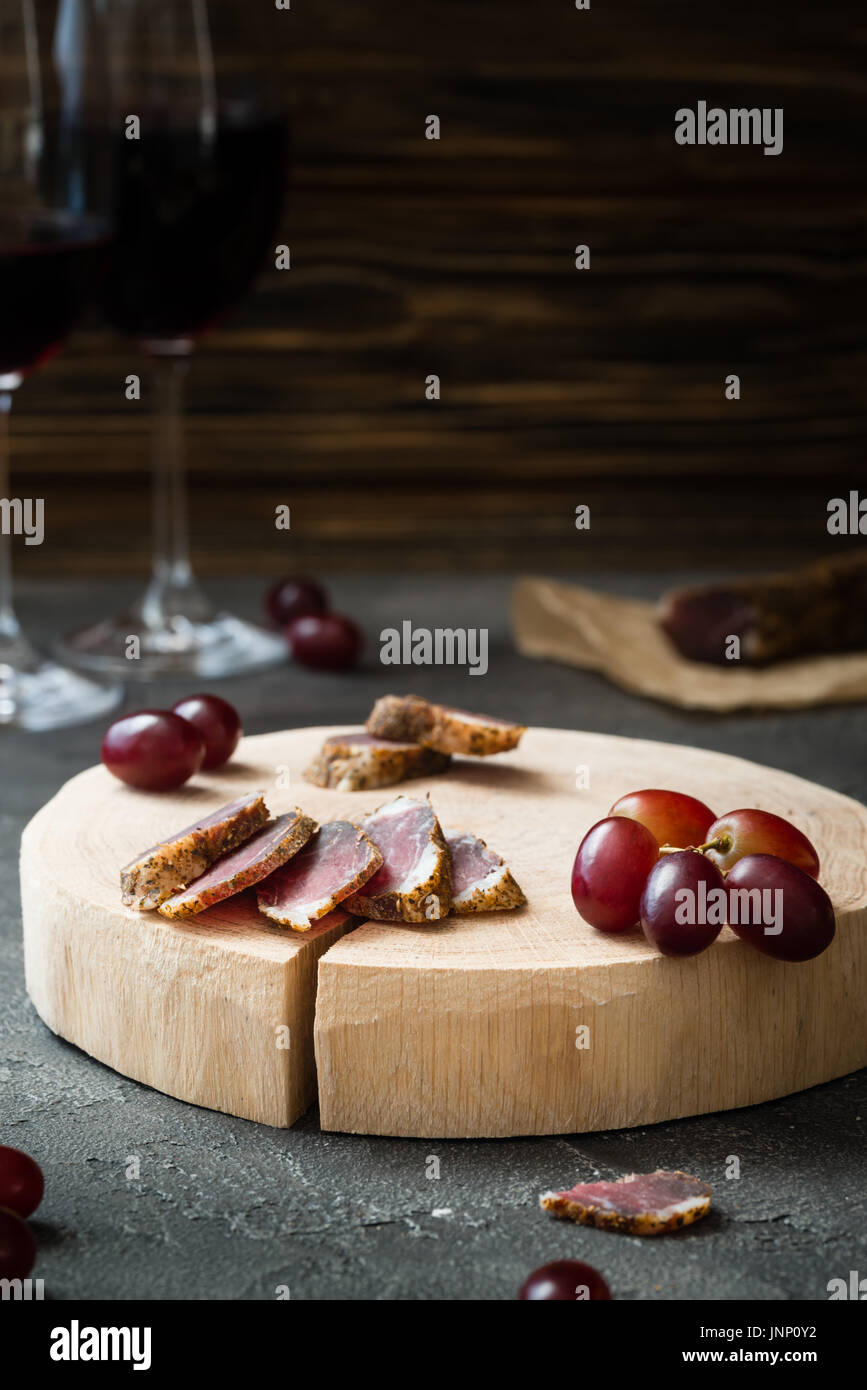 Slices of dried meat on wooden cut, red grapes and two glasses of red wine on dark rustic background Stock Photo