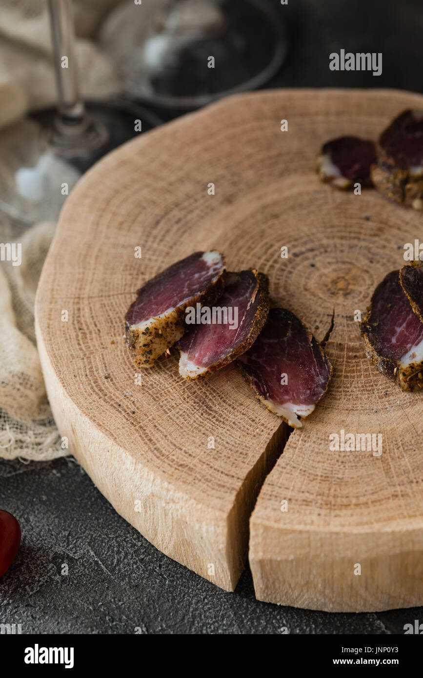Slices of dried meat on wooden cut and two glasses legs on dark rustic background Stock Photo