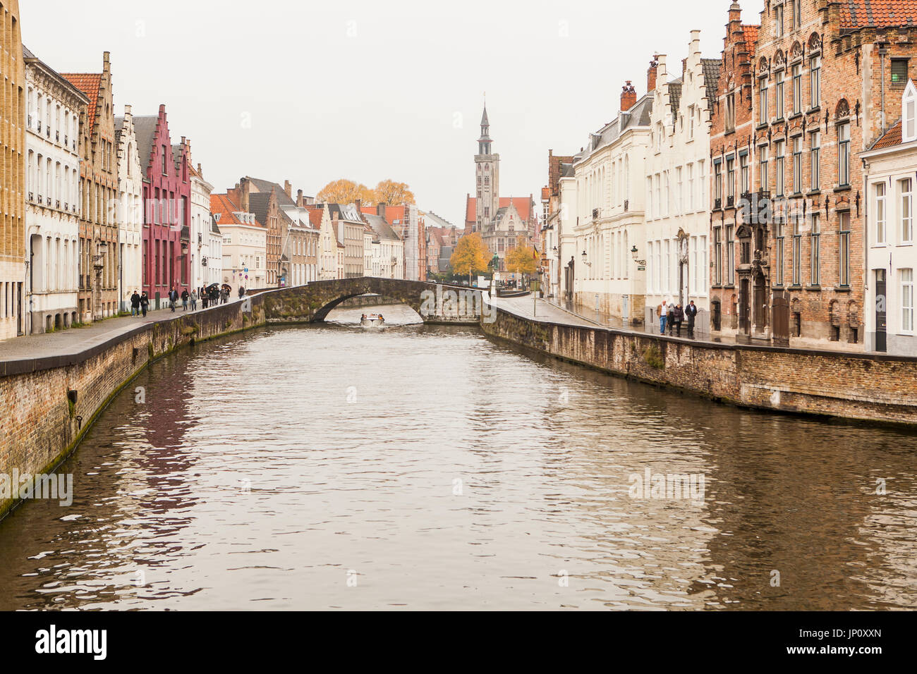 Bruges, Belgium – October 31, 2010: Boat going under the Koningstraat bridge and people walking by the canal in the rain on Spinolarei and Spiegelrei in Bruges, the statue of Van Eyck and the Poortersloge beyond. Bruges is sometimes referred to as the Venice of the North. Stock Photo